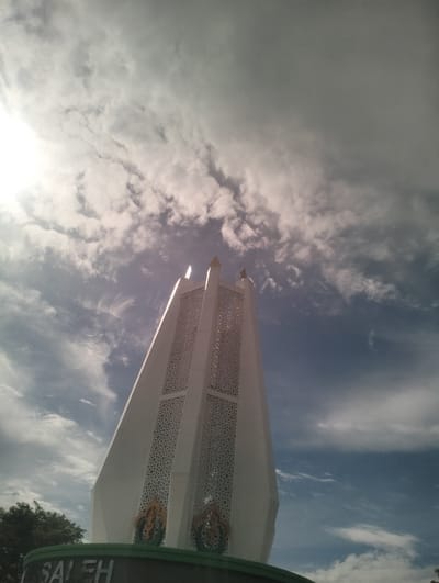 The image shows a low-angle shot of a white, modern monument against a cloudy blue sky. The monument has a tall, tapered structure with intricate geometric patterns on its sides. At the base of the monument, a green sign with white lettering is partially visible, displaying the word "SALEH". The monument appears to be located in an outdoor public space. The sky is filled with both bright sunlight breaking through clouds and darker, more ominous cloud formations. Trees are visible in the lower left corner of the frame. The overall scene suggests a monument or landmark in an urban or semi-urban setting. Based on the provided information, this monument is located in the city of Reuleut Timu, Indonesia.