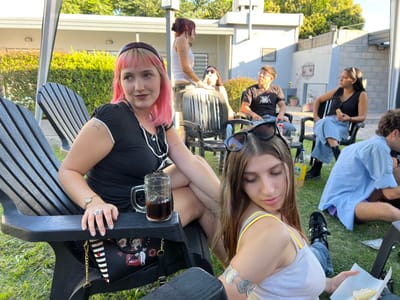 The image depicts an outdoor social gathering of seven young adults in a residential backyard in Quilmes Oeste, Argentina. The scene is illuminated by natural daylight, indicating daytime.
In the immediate foreground, on the left, a female with light pink and blonde hair, styled with bangs and a dark brown headband, is seated in a black Adirondack-style chair. She wears a black short-sleeved top with white trim and buttons, dark lipstick, a ring on her left ring finger, and a star tattoo on her right bicep. Her gaze is directed towards the right side of the frame. A clear glass mug containing a dark brown liquid rests on the chair's armrest beside her. A black handbag, decorated with white stripes and a Betty Boop illustration, hangs from the armrest. To the right, a second female with long brown hair, wearing sunglasses on her head and a white and yellow tank top, is seated on the grass or a low surface. She wears a silver arm cuff on her left bicep and holds a piece of white food in her left hand, looking downwards. Her lower body is visible, showing light blue jeans and black sneakers.
In the mid-ground, a third female with reddish-brown hair and a light-colored tank top is standing behind the pink-haired woman. Further back, a fourth female with dark hair and sunglasses perched on her head is seated in a black patio chair. Adjacent to her, a male with short dark hair is seated in a black patio chair, wearing a black t-shirt with a white graphic design and blue jeans; he is looking upwards and to the right. Next to him, a fifth female with dark hair and a white headband is seated in a black patio chair, wearing a black sleeveless top, light blue wide-leg jeans, and black boots. She holds a piece of food in her hands. On the far right, a sixth male with light-colored, possibly curly hair is seated on the grass, partially facing away from the viewer, wearing a light blue long-sleeved shirt and dark shorts.
The background features a low, light-colored residential building with a flat roof, green hedges, and a concrete perimeter wall. A plastic bottle with a yellow label is visible on a small table situated among the mid-ground group. The ground throughout the visible area is covered in green grass.