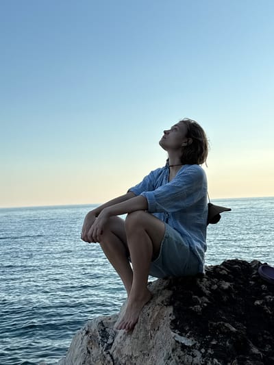 A young person is sitting on a rock by the sea, looking up at the sky. The person is wearing a blue striped shirt and light-colored shorts. They are barefoot. The sea is calm and the sky is clear. The sun is low in the sky, casting a warm glow. The location appears to be coastal, with rocks and water visible. It is not possible to determine the specific location within Donji Katun, Montenegro from the image alone.