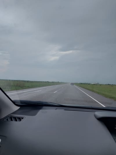 The image, taken from inside a vehicle, shows a desolate highway under a cloudy, overcast sky. The road is wet, suggesting recent rain or damp conditions. The surrounding landscape is flat and consists of green fields with sparse vegetation, extending to the horizon. The highway stretches into the distance, with white lines marking the lanes. A dark-colored car is visible on the road ahead, heading in the same direction. In the far distance, on the right side of the road, there are some buildings under construction, possibly cranes and structures. The interior of the vehicle's dashboard is visible in the foreground, with an air vent on the left. The overall impression is of a journey through a sparsely populated rural area. The scene does not depict any specific landmarks or features that would clearly place it in Novyi Byt, Russia, but the visual elements are consistent with a typical highway in a rural setting.