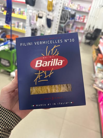 A close-up, top-down shot captures a person's hand holding a box of Barilla brand Filini Vermicelles N°30 pasta. The box is dark blue with prominent white and red text. The pasta itself is visible through a rectangular window at the bottom of the box, appearing as fine, straw-colored strands. The background is blurred, showing shelves stocked with various products, suggesting an indoor retail environment. The overall scene suggests a typical grocery store setting. No specific location in Mayrtup, Russia can be determined from the image.