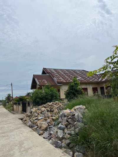 This media file depicts an outdoor scene in Bundar, Indonesia, with a focus on a weathered building and its surroundings. The image is oriented vertically, showing a house with a rusty, corrugated metal roof and cream-colored walls. Several windows with red frames are visible on the side of the house. Lush green grass grows along the side of the building, and a pile of large, grey rocks is situated between the grass and a concrete path. Trees are also present, with some foliage hanging over the roof of the house. In the background, more buildings and palm trees are visible, suggesting a residential or village area. A power line with a single light fixture stretches across the frame. The sky is overcast with clouds, indicating potentially dull or rainy weather. The time of day appears to be daytime. No people are visible in the image, and no text is present. The overall impression is one of a somewhat neglected but functional living space in a tropical or subtropical environment.