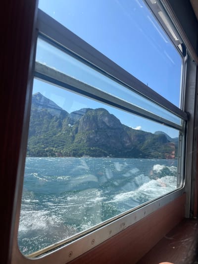 A partially opened window offers a scenic view. The left pane reveals a clear blue sky. Through the right pane, a rugged mountain covered in lush green vegetation dominates the landscape. A body of water, possibly a lake or ocean, is visible in the foreground, with splashes and whitecaps suggesting movement. The window frame is metal, and part of an interior wooden wall is visible. The overall impression is one of travel and natural beauty.