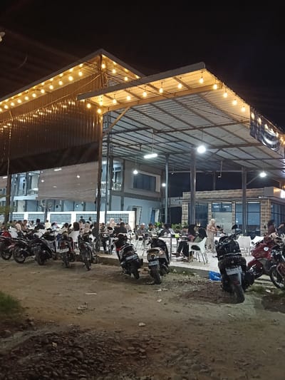 The image captures a nighttime scene at an outdoor dining establishment in Kampongbaro, Indonesia. An unpaved dirt and gravel area in the foreground serves as a parking lot for approximately 15 to 20 motorbikes, primarily scooters, parked in various orientations.

The main subject is a large, open-sided building featuring a high, sloped corrugated metal roof supported by an exposed metal truss framework and columns. Under this roof, an estimated 50 to 70 individuals, primarily adults, are seated at numerous white tables and chairs. They appear to be engaged in dining and social interaction. Some individuals are standing or moving within the establishment.

To the left background, a multi-story building with a dark, possibly corrugated metal facade on its lower section and a horizontal wooden slat design on an upper level is visible. To the right, a single-story building with large glass windows and a textured stone-like facade displays a sign reading "LEWA HOTEL." The entire area is brightly illuminated by numerous warm-toned globe lights strung under the roof structure, complemented by cooler-toned fluorescent or LED lights within the covered space. The sky above is dark, indicating night.