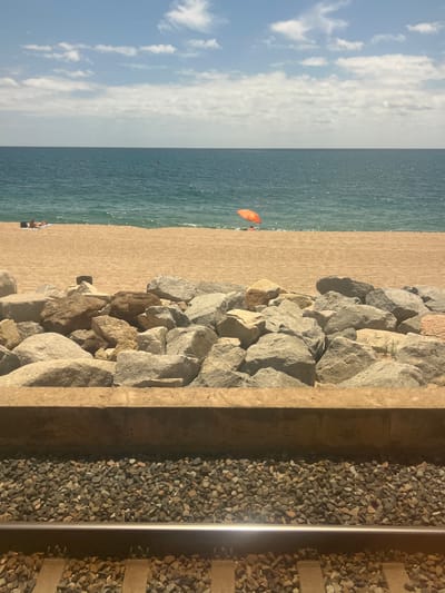 The image captures a vibrant coastal scene, viewed from a slightly elevated position looking across horizontal layers of landscape.
In the immediate foreground, a set of railway tracks runs parallel to the coastline. These tracks feature two shiny metal rails supported by dark wooden sleepers embedded in a bed of dark grey gravel ballast.
Adjacent to the railway, a sturdy concrete wall or barrier separates the tracks from a rugged embankment composed of numerous large, irregular grey rocks. These rocks likely serve as a sea defense or breakwater.
Beyond the rock barrier, a wide expanse of light brown sandy beach stretches towards the sea. Several individuals are scattered across the beach; one person is clearly visible lying down on the sand near the water's edge, while other distant figures suggest more people enjoying the shore.
The beach transitions into the clear, blue-green waters of the sea. Small ripples and sparkles indicate movement and reflections on the water's surface. An identifiable orange object, possibly a small buoy, inflatable, or kite, floats in the sea relatively close to the shore, with other tiny figures further out in the water.
The sky occupies the upper portion of the image, displaying a bright light blue hue adorned with scattered white, wispy clouds. The distinct horizon line separates the calm sea from the sky.
The scene depicts a sunny daytime environment, likely late morning or afternoon, characterized by clear weather conditions. The presence of both a sandy beach and a railway line directly adjacent to the coast strongly suggests a specific type of Mediterranean coastal setting, consistent with the provided location hint of Sant Pol De Mar, Spain. No visible text is present in the image.