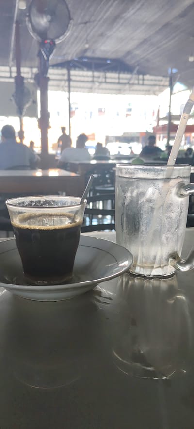 The image presents a scene at an outdoor cafe in Barat Yaman, Indonesia. In the foreground, a small glass cup of dark coffee sits on a white saucer next to a tall glass of cold water with a straw. Behind these, indistinct figures of people occupy tables, suggesting a moderately busy atmosphere. The background is blurred, likely due to the camera's focus on the foreground. Above, a ceiling with hanging fans is visible, indicating an open-air structure. The overall light suggests it is daytime. The table's surface is reflective.