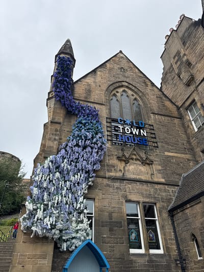 A stone building, possibly a church or historic structure, is adorned with a large, cascading floral installation. The installation features shades of purple, blue, and white flowers, artfully arranged to appear as if they are growing down the side of the building and over a prominent spire. The building itself has a gothic architectural style, with arched windows and stone detailing. A sign that reads "COLD TOWN HOUSE" is mounted on the facade. In the lower left of the image, a person in a red jacket is walking up a set of stone stairs. The sky is overcast. The building is situated in Edinburgh, Scotland, as indicated by the "COLD TOWN HOUSE" sign, which is a well-known establishment in the city.