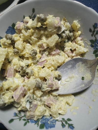 A close-up shot shows a bowl of salad. The salad appears to be a mixture of diced potatoes, hard-boiled eggs, ham, and green peas, all coated in a creamy dressing. A silver spoon rests in the bowl, partially scooping up some of the salad. The bowl itself has a decorative pattern of blue flowers and green leaves on a white background. The overall impression is that of a homemade dish, possibly prepared for a meal. The lighting suggests it could be daytime. No people are visible in the image.