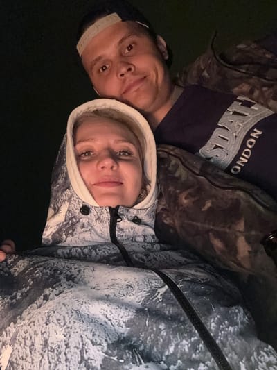 Two individuals are in the image. A man with a baseball cap and a dark t-shirt with "NDON" printed on it is to the left. A woman with blond hair wearing a hooded jacket is to the right. They are posing close together. The environment appears dark. No definitive elements suggest a location in Mayak, Russia.