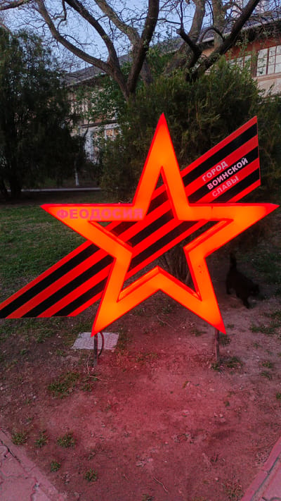 The image shows a lighted star-shaped sign with red-orange illumination, featuring the Russian words "Феодосия" (Feodosiya) and "город воинской славы" (city of military glory). It is positioned on a patch of reddish-brown earth with some grass. A black cat is partially visible behind the sign. In the background, there are trees, bushes, and a building. The overall setting appears to be outdoors, possibly a park or a public area. The location is presumed to be in Feodosiya, which is part of the Орджоникидзевский поселковый совет, Ukraine.