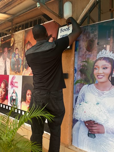 A man is shown from behind, wearing a gray and black short-sleeved shirt and black jeans, affixing a large poster to an outdoor wall. The poster on the right is a portrait of a woman in a wedding dress and tiara. To the left of the wedding portrait are several smaller posters featuring portraits of individuals, including a child and two women, one of whom is holding a balloon shaped like the number "6". Above the posters, there is a corrugated metal roof structure and an outdoor light fixture. A green plant with fronds is in the foreground, partially obscuring the lower portion of the man's legs and the posters. The scene appears to be in Sabojibi, Nigeria.