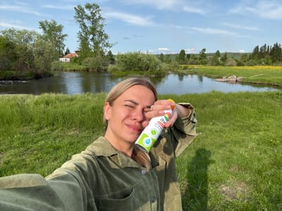 A woman is taking a selfie in a grassy area next to a body of water. She is wearing a green jacket and holding a can of spray in her right hand, which is raised to her face. She is smiling with her eyes closed. The background shows a calm river, lush green grass, trees, and a clear blue sky with scattered clouds. In the distance, there are hints of industrial structures. The location is identified as Nevon, Russia.