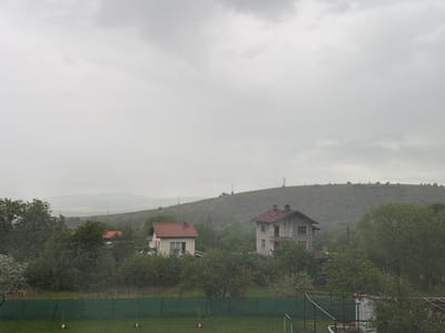 The image shows a landscape in Resilovo, Bulgaria under overcast skies. Two residential houses with red-tiled roofs are visible among greenery. A hill rises in the background, topped with a cell tower. A green fence borders a lawn in the foreground. The overall environment suggests a rural or suburban setting, typical of Resilovo's location in western Bulgaria.