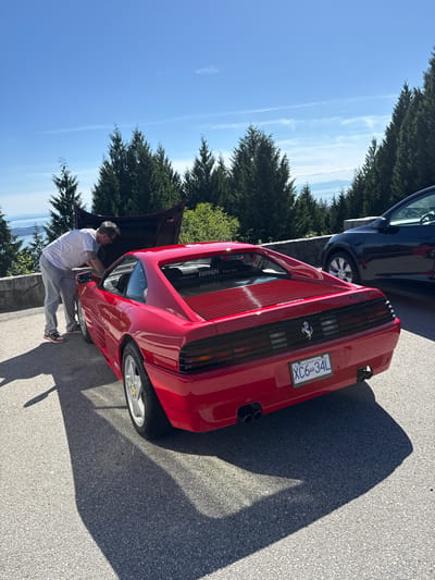 An outdoor image, rotated 90 degrees clockwise, captures a vibrant red Ferrari 348 parked on an asphalt road under a bright, clear blue sky. A man, dressed in a white t-shirt, grey pants, and sneakers, is leaning into the open front compartment (frunk) of the sports car, appearing to inspect its contents.
Behind the Ferrari, a dark-colored vehicle is partially visible. The setting features lush green trees lining the road and hillside, with a scenic backdrop of a large body of water and distant mountains, consistent with the West Vancouver, Canada location. The bright sunlight casts strong shadows on the pavement, suggesting it is midday or early afternoon.
Notable details include the "Ferrari 348" emblem and the "XC6 34L" license plate, which also displays "Beautiful British Columbia." Inside the open frunk, partially visible text includes "Santocar Spa" and "Italia," likely indicating a detail about the car's origin or specifications. The rear of the Ferrari features characteristic slatted vents and dual exhaust pipes, while the open frunk suggests either a quick check or preparation for a journey. The scene conveys a relaxed atmosphere, possibly during a scenic drive or car gathering.