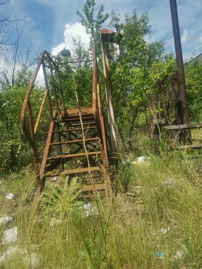 The media file displays an outdoor scene in Verinsko, Bulgaria, characterized by an abandoned, heavily rusted metal structure, likely a staircase or platform, partially consumed by dense vegetation. The structure, appearing corroded with shades of orange and brown rust, stands amidst a profusion of tall green grass and weeds in the foreground, with lush green trees and bushes forming a vibrant backdrop. Some white blossoms are visible among the foliage.
The sky above is bright blue with scattered white, fluffy clouds, indicating a sunny day. A notable detail is a large bird's nest perched high within the framework of the rusted metal structure, with a bird clearly visible resting on it. This highlights nature's adaptation and reclamation of the abandoned site. Scattered pieces of plastic debris, including white and blue items, are visible among the tall grass at the base of the structure, indicating human impact and neglect. No people are present, and there is no visible text in the image. The scene evokes a sense of decay and natural resurgence.