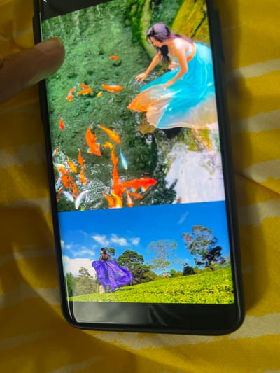 A smartphone, held by a person's finger, rests on a yellow and white striped fabric. The phone's screen displays two vibrant vertical images side-by-side, viewed horizontally.

The image on the left shows a woman in a flowing, translucent blue and orange gown. She has long dark hair and is leaning over a body of water, possibly a pond or stream. Numerous colorful koi fish, predominantly orange with some white and light blue, swim actively beneath the water's surface amidst green aquatic vegetation. The setting appears to be an outdoor, lush green environment. The woman's pose suggests an interaction with the fish, perhaps feeding or admiring them.

The image on the right features a different woman wearing a striking, flowing purple gown. She is standing amidst a vast, undulating landscape of bright green, manicured vegetation, strongly resembling a tea plantation. The sky above is a clear blue with scattered white clouds, and a few trees are visible in the distance. The woman is positioned with her back partially to the viewer, showcasing the dramatic effect of her purple dress against the verdant backdrop.

Both images depict bright, sunny daytime outdoor scenes. The overall composition suggests a display of scenic beauty and elegant portraiture, potentially from Ngasinan, Indonesia, as indicated by the context. No visible text is present in the images or on the phone screen.