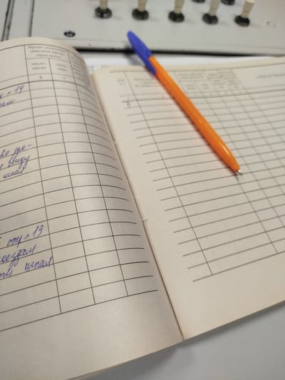 A close-up shot captures an open notebook with a grid of lines and handwritten entries in blue ink. The notebook is lying on a light-colored surface, possibly a desk. A bright orange and blue ballpoint pen rests diagonally across the notebook's pages, with its tip pointing towards the right side of the image. The notebook's left page has headings like "Время окончания действия", "число", and "месяц", with a handwritten "8" under "месяц". Further down, entries include "+14", and what appears to be "школы". The right page is mostly blank grid lines, with a faint blue mark near the top. In the background, out of focus, are some beige and black objects with circular bases and vertical stalks, suggesting some kind of electronic equipment or control panel. The scene is devoid of any specific environmental context that would place it in Krasny Gulyai, Russia.