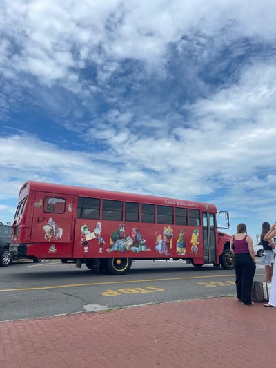 The image shows a bright red bus with cartoon characters painted on its side, parked on a paved road. The text "Camp Jabberwocky" is visible on the side of the bus. In the foreground, three women are standing on a red brick sidewalk, facing away from the camera. The bus is likely in Oak Bluffs, Massachusetts, a town known for its colorful gingerbread cottages and the historic Flying Horses Carousel, as well as Camp Jabberwocky, a camp for people with disabilities. The sky is blue with scattered white clouds.
