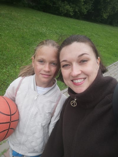 In this selfie-style photo, two females, a woman and a young girl, are pictured outdoors. The woman is on the right, and the girl is on the left. The woman has dark hair and is smiling broadly, revealing white teeth. She is wearing a dark brown sweater with a high collar that has a silver peace sign pendant attached to the zipper. Her eyes are open and she is looking directly at the camera.
The young girl has blonde hair pulled back in a ponytail and is also looking at the camera. She has glitter makeup on her cheeks and nose, and is wearing a white quilted jacket over a white shirt. She is holding an orange basketball in her left arm. The girl appears to be wearing a light pink backpack with pink straps.
The background consists of a grassy area with small white flowers, suggesting a park or a lawn. Behind the grass, there is a dark green wooded area, likely trees. A paved walkway is visible to the right of the woman. The lighting suggests it is daytime, and the greenery indicates warm weather, likely summer. The overall mood appears happy and casual, as if they are enjoying an outing together, perhaps a game of basketball. The location context is Tosno, Russia. There is no visible text in the image.
