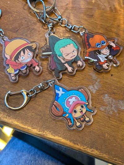 Four One Piece character keychains are displayed on a wooden surface. The keychains feature Monkey D. Luffy wearing his straw hat, Roronoa Zoro with green hair, Portgas D. Ace in his signature hat and outfit, and Tony Tony Chopper in his hat and blue coat. The keychains are made of clear acrylic and are attached to metal keyrings and clasps. The wooden surface appears to be a table with a visible grain and some dust or debris. The lighting suggests an indoor environment. This image does not provide any information that can be used to determine its location in Candirejo, Indonesia.