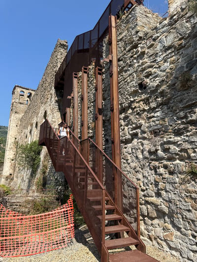 A woman walks down a rusty metal staircase attached to a stone building in Dolceacqua, Italy. The building appears to be old and possibly in disrepair, with parts of the stone wall crumbling and overgrown with vegetation. A bright orange plastic safety fence is visible in the foreground. The sky is clear and blue, indicating a sunny day. The staircase and its supporting structure are made of dark, weathered metal, with a grated surface for the steps and railings. The woman is wearing a white t-shirt and dark pants, carrying a backpack. The stone wall itself is constructed from irregular-shaped stones of varying sizes and colors.