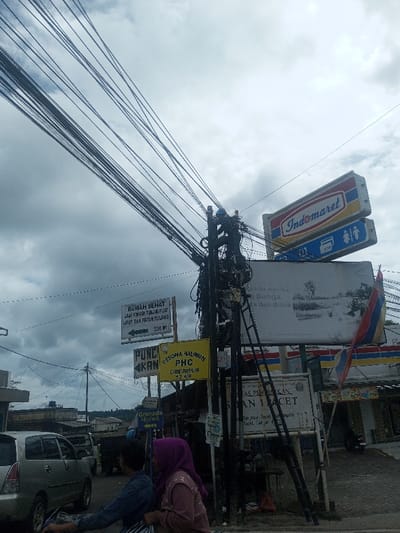 The image presents a street scene in Sukanagalih, Indonesia, under a cloudy sky. A densely wired utility pole dominates the central frame, supporting numerous black electrical and communication cables that extend diagonally upwards to the left. A black multi-section ladder leans against the right side of this pole.
To the pole's right, an "Indomaret" sign is prominently displayed above a sign indicating ATM and restroom facilities. A large billboard, partially obscured by the pole, features a landscape image and some text. A red and blue striped flag is affixed near an Indomaret store entrance, visible on the far right.
To the pole's left, a white directional sign points left, stating "RUMAH SEHAT Ahli Hancur Tulang & Patah Tulang 200 M." Below it, a yellow sign reads "PESONA HALIMUN PHC CIBEREUM 2 KM." A partially visible white sign labeled "PUNCAK KAN" is further to the left, with a "Granada SkyPark" sign below it. Near the base of the ladder, a white sign for "DINAS PENDIDIKAN SMAN 1 PACET" is visible.
In the lower foreground, two individuals are riding a motorcycle, moving from left to right. The person in front wears a dark blue long-sleeved shirt. The passenger behind wears a purple hijab and a light brown top. Their faces are largely obscured. Behind them, a silver minivan, likely a Toyota Innova, is partially visible, moving in the opposite direction. Additional buildings and vehicles are present in the background.