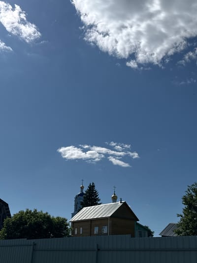 The image depicts a clear blue sky with scattered clouds above a rural or small-town landscape. In the foreground, a light gray corrugated metal fence runs horizontally across the lower portion of the frame. Behind the fence, a wooden building with a metallic, sloped roof is visible, featuring several windows. To the left of this building, a church with a blue dome and a golden cross is partially obscured by a dark green conifer tree. Further to the right, another building with a dark, sloped roof and a glimpse of green siding can be seen, also with trees in the background. The scene is set in Новосёлки, Russia.