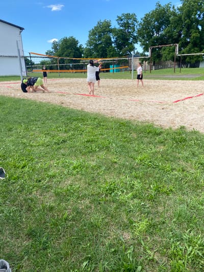 A group of young people are playing beach volleyball on a sandy court next to a grassy area. The scene is outdoors under a clear blue sky with a few white clouds. The sun appears to be shining, suggesting a bright, warm day, likely during daytime hours. Trees are visible in the background, indicating a park or recreational area.

There are at least four individuals visible on the court. Two people are on one side of the net, one is in the process of hitting a volleyball, and the other is in a defensive stance. On the other side of the net, two other individuals are present. One is standing, seemingly observing the game, and another is in a crouched position. They are all dressed in athletic wear, suitable for playing sports.

The court has distinct red boundary lines marking the playing area. A volleyball net stretches across the center of the court. Adjacent to the sand is a well-maintained grassy area, suggesting the volleyball court is part of a larger outdoor facility. In the far background, there are some structures that appear to be part of the surrounding environment, possibly fencing or buildings.

The activity is clearly a game of beach volleyball, with players actively engaged in the sport. The mood seems to be casual and recreational, with the focus on the game. No visible text is present in the image. The location context provided is Mostovskoy, Russia.