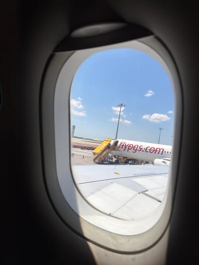 The media file shows a view from an airplane window of a Pegasus Airlines aircraft on the tarmac. The aircraft is white with red lettering that reads "flypgs.com". A yellow airstair is connected to the aircraft's door. In the background, there are airport lights and a bright blue sky with scattered white clouds, suggesting it is daytime with fair weather. The view is framed by the curved interior of the airplane window. The scene appears to be at an airport, possibly in Pendik, Turkey, given the context. The image conveys a sense of travel and departure.