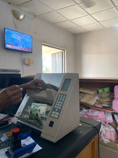 The image depicts an indoor scene, likely an office or a home, in Jere North, Nigeria. The primary subjects are a person's hands interacting with a cash counting or deposit machine. The machine, a light gray, has a digital display and a keypad with numerous circular buttons. The drawer of the machine is open, revealing stacks of currency, some of which appear to be Nigerian Naira.
To the left of the machine, a flat-screen television is mounted on the wall, displaying a news program with the text "2022 ELECTION COUNTDOWN" and other smaller text at the bottom. The television is positioned near a light fixture on the wall.
In the background, a bookshelf is visible, filled with what appear to be files, papers, and possibly some pink folders. The overall setting suggests a workspace with a mix of modern equipment and paper-based storage. The lighting in the scene is ambient, and it's difficult to ascertain the exact time of day or weather conditions. The focus of the image is the interaction with the cash handling machine, with the person's hands actively engaged in its operation. The emotions conveyed are neutral, focusing on the task at hand.