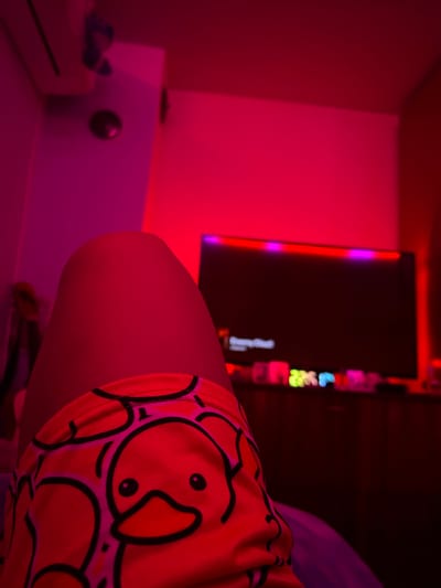 The image depicts an indoor room saturated in strong red and magenta ambient lighting, located in Debelt, Bulgaria. In the lower foreground, a person's bare upper leg is visible, clad in orange shorts adorned with a repeating pattern of black-outlined, white-faced cartoon ducks featuring orange beaks. The leg is angled from the lower left towards the center. In the background, a large, dark flat-screen television is visible, its top edge illuminated by an LED strip displaying alternating red and purple lights. Below the television, a surface holds several small, brightly illuminated objects, some appearing green, blue, and pink. To the upper left, a white wall-mounted unit, consistent with an air conditioner, is present, with a dark, circular object mounted on the wall directly beneath it.