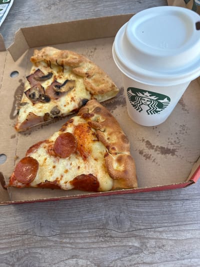 A high-angle shot captures two slices of pizza and a Starbucks coffee cup inside a red and brown pizza box. The location is Mougins, France, as inferred from the presence of a Starbucks (though this is not directly visible in the image). One pizza slice has pepperoni toppings, while the other has mushrooms and ham. The pizza box appears slightly stained with grease. The background is a grey, wood-like surface. There are no people visible in the frame. The scene appears to depict a casual meal setting.