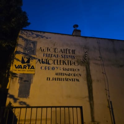 The image is a medium-shot, eye-level, outdoor photograph taken at dusk or night, featuring the exterior wall of a building in Piešťany, Slovakia. The wall is a dull yellow-beige color, showing signs of age and wear, with vertical streaks of dark discoloration and a shadow from a tree on the left side.

Dominating the center of the wall are painted signs. The top sign, in large, dark blue, blocky serif letters, reads "AUTO BATÉRIE". Below this, in smaller, dark blue serif letters, is "PREDAJ-SERVIS". Further down, in larger, dark blue, stylized serif letters, is "AUTOELEKTRO". Underneath "AUTOELEKTRO", in dark blue serif letters, is "OPRAVY : ŠTARTÉROV", followed by "ALTERNÁTOROV" and "EL.PRÍSLUŠENSTVA". To the right of the main text, written vertically with an arrow pointing left, is the word "SERVIS" in black, handwritten-style lettering.

On the left side of the wall, a yellow rectangular sign with a black logo is prominently displayed. The logo consists of two V-shapes forming a stylized "M". Below the logo, in bold black letters, is "VARTA", and beneath that, in smaller black letters, reads "THE BATTERY EXPERTS". Behind the Varta sign, a vertical dark blue rectangle with an "A" painted inside is visible, suggesting a previous or overlapping sign.

At the top center of the wall, a brick chimney with a metal cap protrudes upwards against a dark blue sky.

In the foreground, a black metal railing with vertical bars runs horizontally across the bottom of the image, obscuring the lower part of the wall and the ground. The scene is illuminated by artificial light, casting shadows and highlighting the textures of the wall.