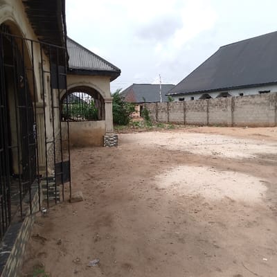 The image shows an empty dirt lot, located at 5.953 latitude, 5.705 longitude, in the city of Adanisievbe, Nigeria. The lot is bordered by a low, concrete wall to the right and a house to the left. The house has a black metal roof and a white, plaster facade. The entrance to the house is covered by a black metal gate. There are bushes and trees visible to the right and behind the house. The lot is mostly dirt, with some patches of grass and debris. There are no people or animals visible in the scene. The sky is overcast.