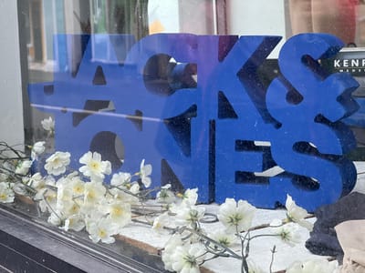 The image shows a close-up, eye-level, outdoor shot of a blue, 3D sign that spells out "#JACKJONES" in a sans-serif font. The sign is illuminated by natural light and has a slightly textured surface with small dark speckles. In the foreground, there is a decorative arrangement of artificial white flowers with yellow centers, draped across the bottom of the frame. The flowers are mostly out of focus but clearly visible. Behind the sign, blurred reflections of buildings and people can be seen, suggesting the sign is displayed in a shop window. The overall scene appears to be a commercial storefront. The location is the town of Donegal, Ireland.