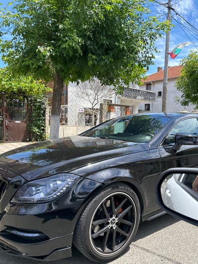 A black Mercedes-Benz sedan is parked on a street in Aheloy, Bulgaria. The car is facing the right of the frame, with its front passenger side and wheel prominently displayed. The vehicle has a sleek, modern design, with LED headlights and a sporty black rim. The tire has "NEXEN" written on it. A side mirror reflects a bit of the surroundings, including some greenery.

Behind the car, a residential building with a red-tiled roof and light-colored walls is visible. The architecture suggests a European setting. A Bulgarian flag, with its white, green, and red stripes, is flying from a utility pole in the background, adding a clear location cue. Lush green trees with dense foliage flank the street, providing shade and a natural element to the scene. The sky is bright blue with some scattered clouds, indicating a clear and sunny day. The time appears to be daytime. No people are visible in the image. The overall scene is calm and typical of a residential street in a town.