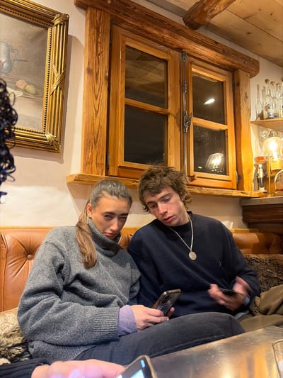 An indoor scene depicts two young adults seated on a brown tufted leather couch, engaged with smartphones, within a rustic establishment consistent with an alpine lodge or restaurant in La Clusaz, France.

The individual on the left is a female with long brown hair tied in a ponytail, wearing a grey turtleneck sweater over a light purple long-sleeved shirt, and dark pants. She holds a dark smartphone horizontally with both hands, looking towards the camera with her left eye partially closed.

The individual on the right is a male with messy light brown hair, wearing a dark blue crew-neck sweater and dark pants. He has a silver-colored chain necklace with a circular pendant. He holds a dark smartphone in his right hand, looking downwards at the screen.

In the foreground, the hands of a third person are visible, also holding a dark smartphone over a reflective surface.

The background features a prominent wooden-framed window with multiple panes and antique metal latches, flanked by thick wooden beams. To the left of the window, a framed painting in an ornate gold frame depicts a still life. To the right, a wooden shelf unit holds several clear glass bottles, and a decorative lamp with a visible filament bulb provides warm ambient light. The interior exhibits extensive use of natural wood, including a visible ceiling beam, reinforcing a traditional alpine chalet aesthetic.