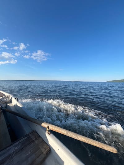This is a tilted, eye-level shot from a boat moving on the water, with the horizon line running vertically. The scene is split between a bright blue sky and the dark, rippling water. A white boat is partially visible on the right, with a wooden oar or pole extending diagonally across the frame. The water churns with white foam and spray, indicating movement. Scattered white clouds dot the sky. The overall impression is of a sunny day, likely midday, with calm but moving water. The image conveys a sense of travel and the open air. No people are visible. The location is Siukterka, Russia.