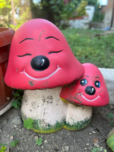 A pair of ceramic mushroom garden decorations, painted bright pink with cartoonish faces, are positioned outdoors on gravel. The larger mushroom, on the left, has closed eyes and a wide smile. The smaller mushroom, nestled to its right, has open blue eyes and a smile. Both are speckled with dirt. The background shows blurred greenery and what appears to be the side of a building, suggesting a garden setting. The location is Belashtitsa, Bulgaria.