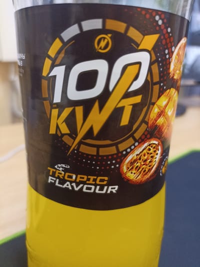 A close-up, eye-level, horizontal shot shows a yellow beverage bottle with a black label that prominently displays the number "100" in white with a yellow outline, followed by "KWT" in large yellow stylized lettering. The label also features a circular design with segmented orange, yellow, and gray accents, and the words "TROPIC FLAVOUR" in white lettering at the bottom. Two halved passion fruits are depicted on the right side of the label. The bottle is positioned on a surface that appears to be a desk or table, with a black mousepad visible in the foreground and a blurred background that includes what looks like a computer monitor and other office items. The lighting suggests an indoor setting, possibly a home or office environment. There is no indication of the city of Shakhunya, Russia.