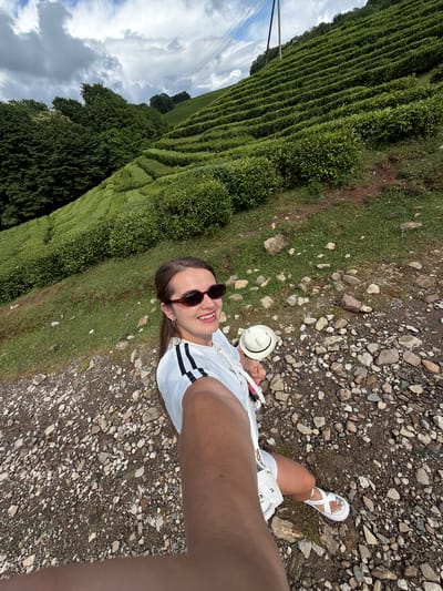 A young woman with brown hair and sunglasses is taking a selfie on a gravel path. She is wearing a white t-shirt with black stripes on the sleeves, white shorts, and white sandals. She is holding a white hat in her left hand. The path is on the side of a steep hill covered in rows of green tea bushes. Trees are visible on the left side of the hill and in the distance. The sky is cloudy. The scene is located in Izmaylovka, Russia, likely a tea plantation given the prominent tea bushes.
