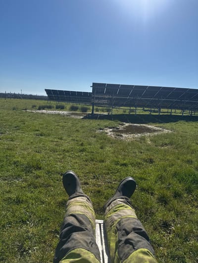 The image depicts a first-person perspective view from a seated or reclined position within a solar farm in a rural area of Eastleigh, United Kingdom, under a clear, sunny sky. The subject's lower legs and feet are visible in the foreground, wearing dark rubber or safety boots and dark trousers with horizontal bright yellow/lime green reflective bands, consistent with protective workwear. The legs rest on a metallic, ribbed surface. The ground ahead consists of a green grassy field with several distinct patches of mud and standing water. In the midground and background, multiple rows of large, dark solar panels, mounted on metal support structures, extend across the landscape. The panels are angled upwards, facing away from the camera's perspective. The sky is bright blue with visible direct sunlight originating from the upper right quadrant, casting strong illumination over the scene. A faint contrail is discernible in the upper left sky. A distant treeline and fence are visible along the horizon.
