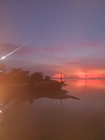 This image captures a tranquil coastal scene in Baan Wok Tum, Thailand, at twilight. The sky transitions from a deep purple at the top to a vibrant gradient of pinks, oranges, and reds near the horizon, reflecting in the calm water. A silhouette of a boat is positioned on the shoreline, its reflection mirrored on the water's surface. The shoreline is visible in the foreground, and some tree are on the shore.