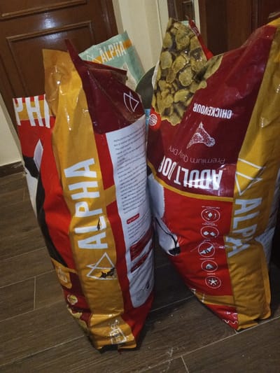 The image depicts an indoor scene in Madinaty, Egypt, featuring multiple bags of pet food. In the foreground, two large, vertically oriented bags of "ALPHA" brand dog food dominate the frame. The left bag, primarily red and yellow/gold, displays "ALPHA" prominently along with text regarding "premium dog food" and a white dog illustration. This bag exhibits a noticeable tear or breach on its lower side, potentially exposing contents. The right bag, also red and yellow/gold, is labeled "ADULT DOG" and "CHICKENOUR PREMIUM DRY," and shows dog food kibble visible at its open top. It also features a dog illustration and several illustrative icons detailing product benefits. In the background, partially obscured, a third, lighter green/blue "ALPHA" branded bag is visible. The floor is tiled with a light brown and grey wood-effect pattern. A dark brown wooden door with panels is visible in the background on the left side. The scene is static, with no individuals or active events present, indicating a storage or domestic setting within the city.