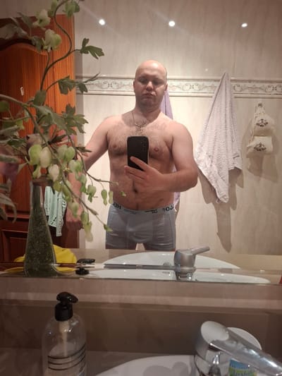 A man is taking a mirror selfie in a bathroom. The man is bald and shirtless, wearing grey underwear. He is holding a black smartphone in his right hand. A gold chain is visible around his neck. The bathroom has beige tiled walls with decorative borders. A white towel hangs on the wall. A sink, faucet, and soap dispenser are visible in the mirror. A vase with greenery stands in front of the mirror. Based on the user prompt, the location is presumed to be in a bathroom somewhere in Xirivella, Spain.