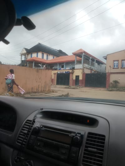 The image, captured from inside a vehicle, shows a street scene in Ifo, Nigeria. In the foreground, the dashboard of the car is visible. To the left of the dashboard, a person is seen walking along a dirt road. The person is carrying a bag over their left shoulder and holding a folded piece of cloth in their right hand. The person is wearing a white t-shirt and a patterned skirt.
The background features several multi-story buildings with red tiled roofs. One building on the right has a light brown facade and white-framed windows. A large black metal gate is visible, separating the buildings from the street. Overhead, power lines are strung across a cloudy sky. The overall environment suggests a residential or commercial area within the city.