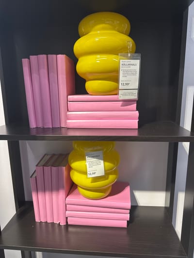 This image shows two black shelves filled with decorative items. On the top shelf, a large, yellow, bulbous vase sits on a stack of pink books. A price tag is attached to the vase. Next to the vase, a stack of pink books is visible. The bottom shelf also contains a similar yellow vase, price tag, and a stack of pink books. The background appears to be a white wall. The scene is indoors, possibly in a retail store. The city of Siero, Spain is not depicted in this image.