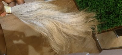 The image displays a close-up view of a hand holding a bundle of light-colored hair, consistent with a setting in Okere-Urhobo, Nigeria. A human hand, exhibiting a medium-dark skin tone, grasps the secured end of a hair bundle. The hair, approximately 15-20 centimeters in length and appearing to be synthetic or processed human hair, is predominantly platinum blonde to light golden blonde, with some darker strands interspersed. The hair is secured at the point of grasp by a light, possibly white or cream-colored, elastic band or tape. The strands are mostly straight but exhibit a slight natural wave or curl towards the ends. The background on the lower left consists of a smooth, light brown surface, possibly a wooden table or floor, where distinct shadows from the hand and hair are visible. The upper right portion of the background features a section of vibrant green, textured material resembling artificial turf or short-pile carpet, situated next to another light brown, planar surface. No other individuals are present in the frame.