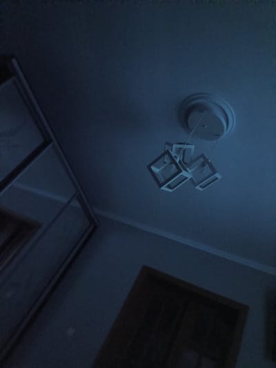 The image depicts an interior scene with a dark blue hue, likely taken at night or in a dimly lit room. In the upper portion of the frame, a modern chandelier with three cube-shaped fixtures hangs from the ceiling. The ceiling itself features a decorative medallion around the light fixture's base. To the left, a large wardrobe or closet with mirrored doors is visible, reflecting the dim light. The lower part of the image shows a doorway with a dark wooden door. The overall environment suggests a residential setting. No people are present in the image. This scene is located in Striletskyi Kut, Ukraine.