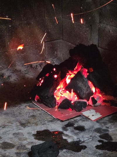 The image displays a contained charcoal fire within a dark, brick or concrete block structure, consistent with a grill or fireplace, located in Villa San Andrés, Argentina. A central mound of dark charcoal embers emits intense red and orange light, actively projecting numerous bright, linear sparks upwards and outwards into the dark ambient space. The burning embers rest on two partially scorched red cardboard or thin board pieces. The immediate foreground shows a light grey, stained concrete or stone floor, exhibiting dark, irregular residue and a distinct unlit charcoal lump in the lower portion. The background consists of dark grey rectangular block masonry. No individuals are present in the scene, which is illuminated solely by the burning charcoal.
